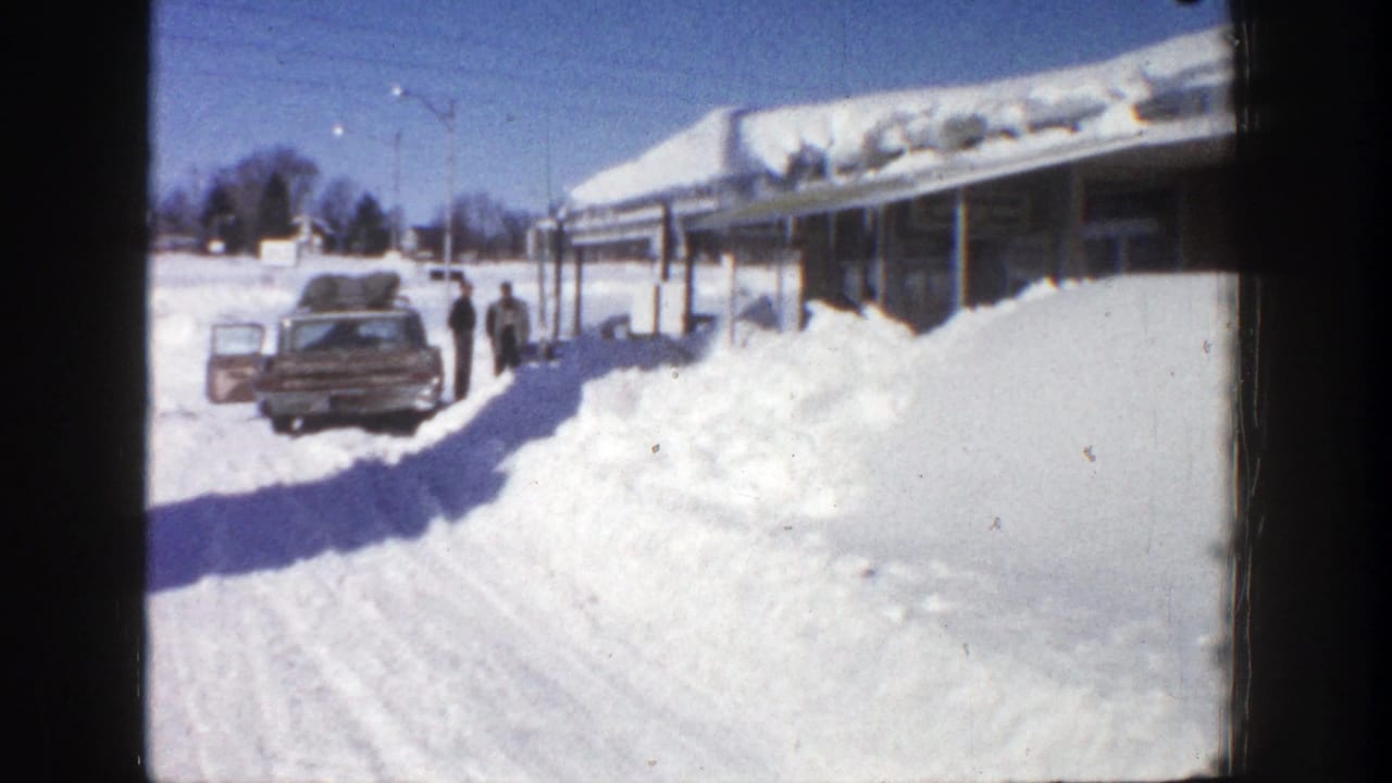 Parked car beside high snowbanks and a snow-covered building, with two people standing nearby.
