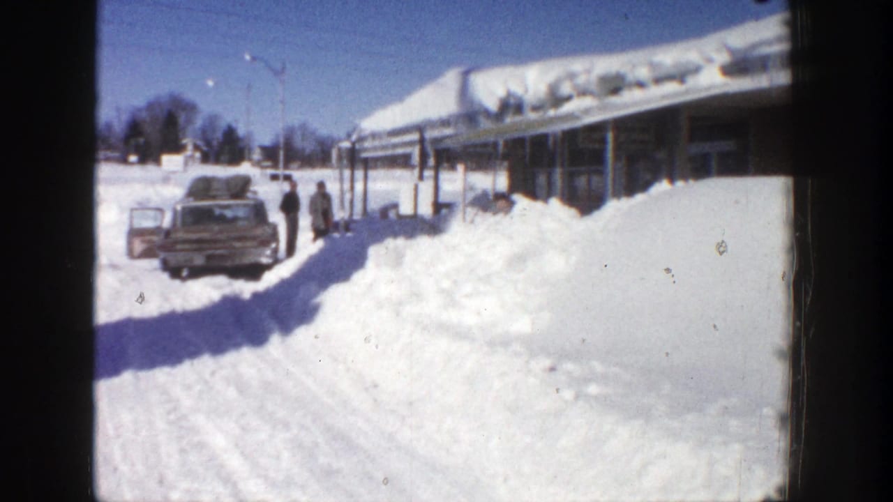 Car parked by a snowy roadside with people near a long building under a heavy snow-covered roof.
