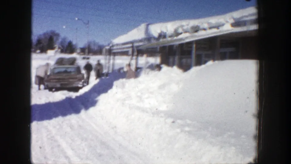 1961 Aspen winter frame with a mid-century car, pedestrians, deep roadside snow, and a low building under a snow-heavy roof.