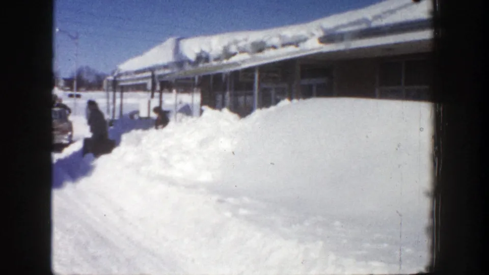 Aspen winter home-movie frame showing a bundled person walking past deep snow, with a car and snow-covered building behind.