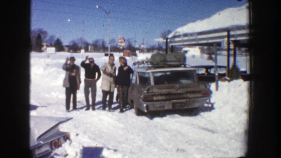 One adult waves toward the camera as the family group stands beside a station wagon in snowy Aspen, Colorado.