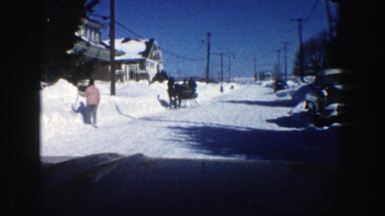 Snow-covered residential street with a distant horse-drawn sleigh and a person shoveling at left.