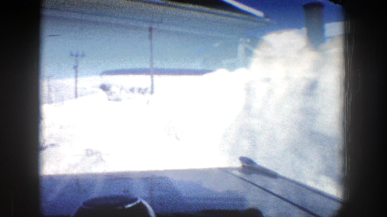 View through a vehicle window toward high snowbanks beside a snowy road.