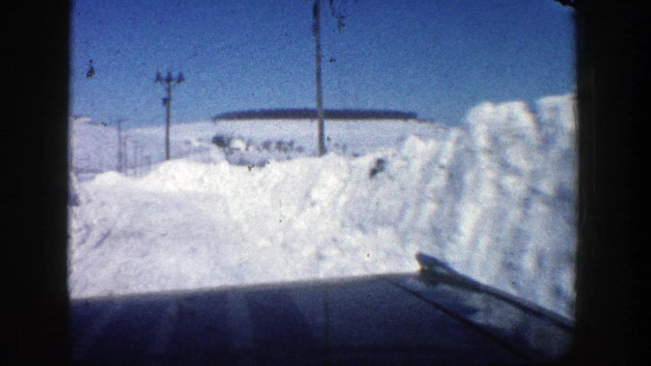 Snowy road bordered by tall snowbanks with utility poles and a distant house.