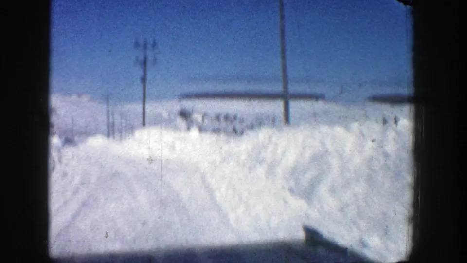 1961 Aspen winter frame with high roadside snowbanks, utility poles, and distant snow-covered ground seen from a moving vehicle.