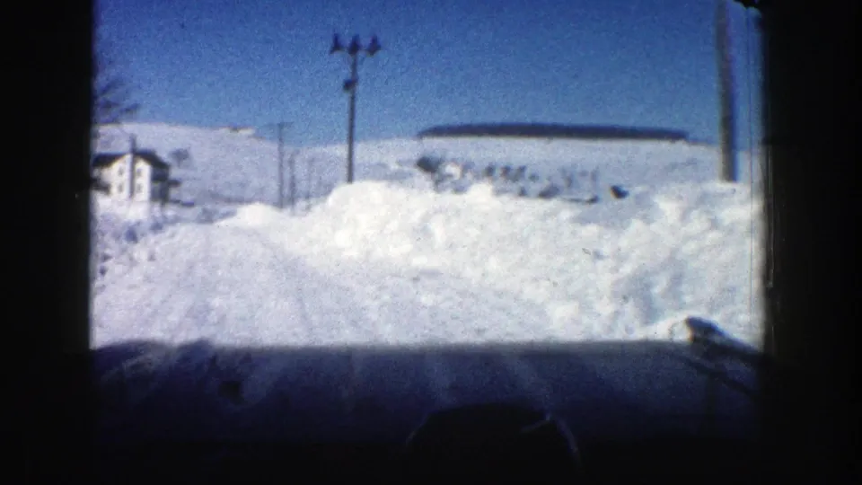 Aspen, Colorado 1961 passenger-view frame showing a snow-covered road, high snowbanks, utility poles, and a white house ahead.