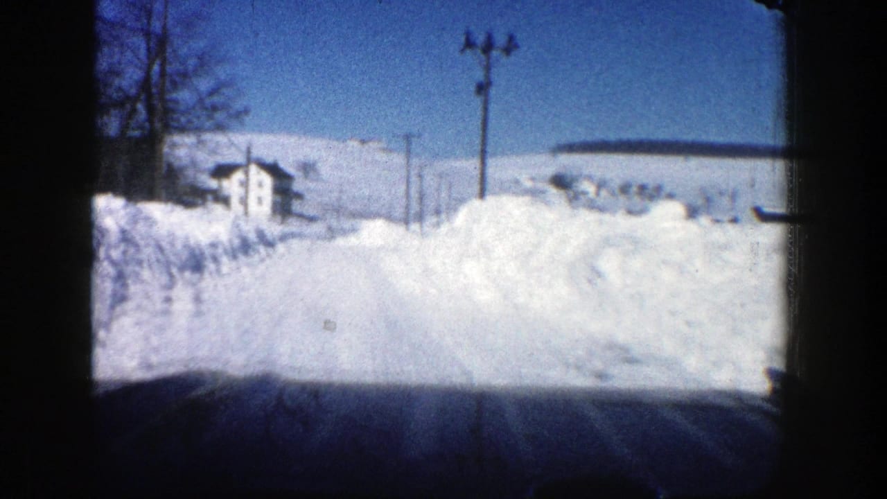 Snow-covered road seen from inside a car, with high snowbanks, utility poles, bare trees, and a distant house.