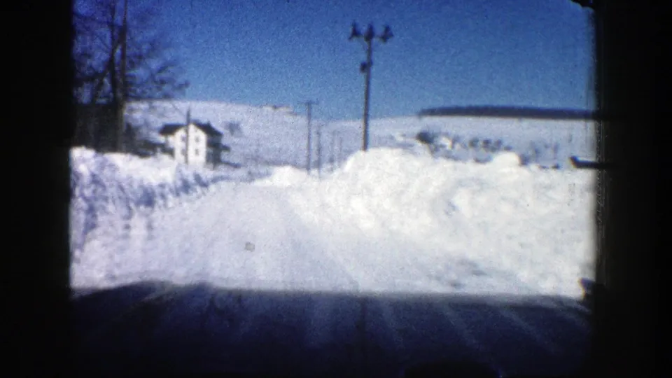 1961 Aspen, Colorado car-window view of a snow-covered rural road with deep banks, utility poles, bare trees, and a small distant house.