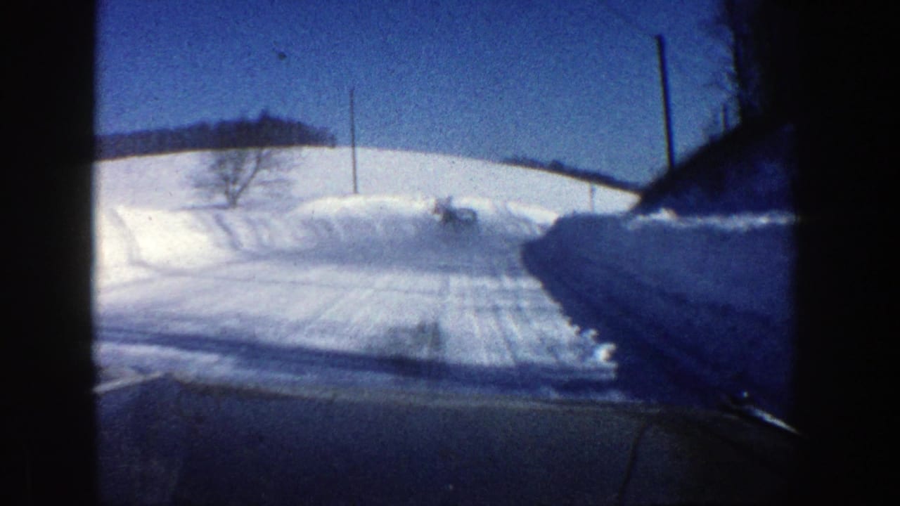 Plowed snowy road curves ahead between tall snowbanks, with a utility pole and low hills under blue sky.