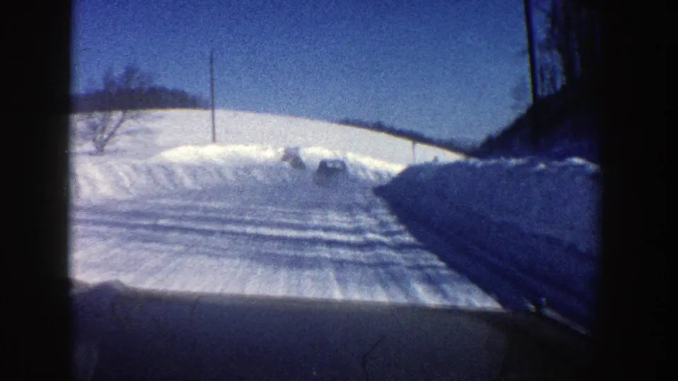 1961 Aspen, Colorado winter road seen from a moving car, with compacted snow tracks, high banks, utility poles, and bare trees.