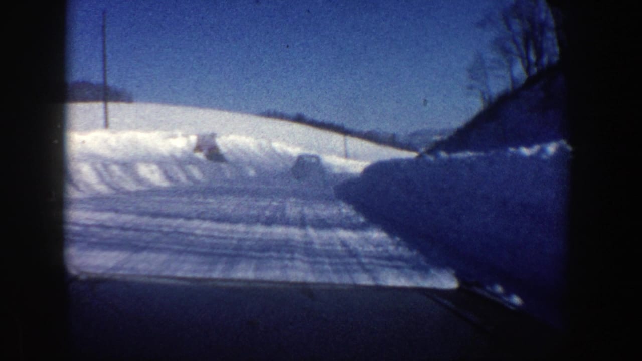 Snow-covered road seen through a car window with high snowbanks and a vehicle ahead.
