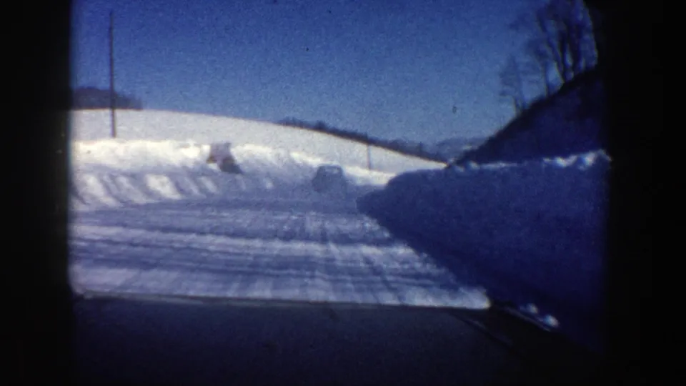 1961 Aspen-area winter road seen from inside a moving car, with plowed snowbanks, blue sky, and a vehicle in the distance.