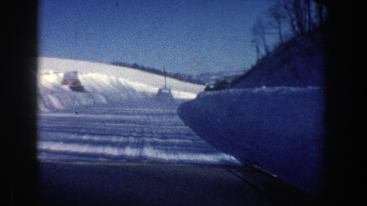 Plowed snowy roadway with snowbanks, a vehicle ahead, and bare trees on a hillside.