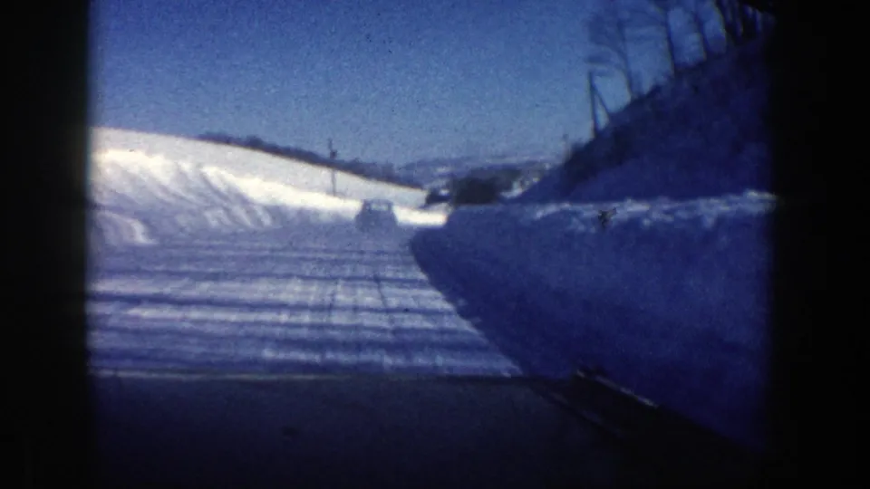 In-car 8mm view of a snowy Aspen-area road in 1961, with plowed banks and a dark vehicle form beyond the curve.