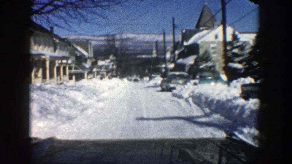 1961 Aspen residential street with deep snowbanks, parked cars, houses, and bare trees seen from a moving vehicle.
