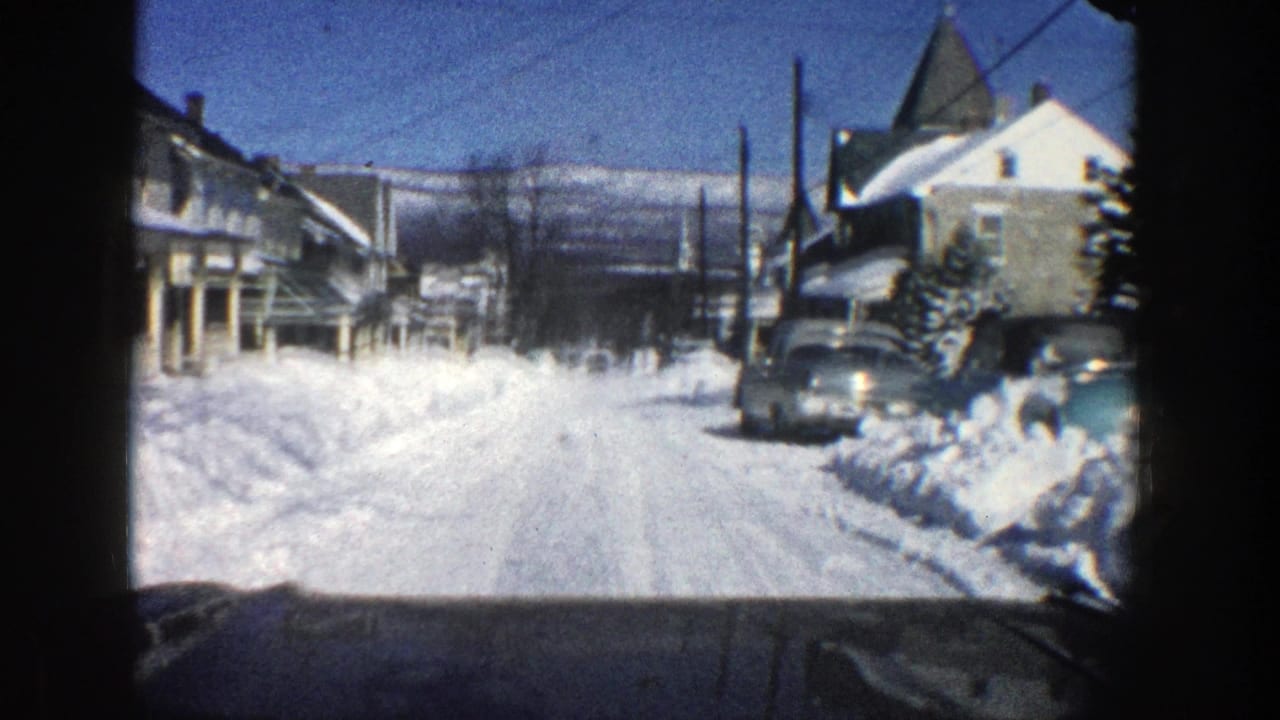 Snowy neighborhood road with parked car and houses under a blue sky.