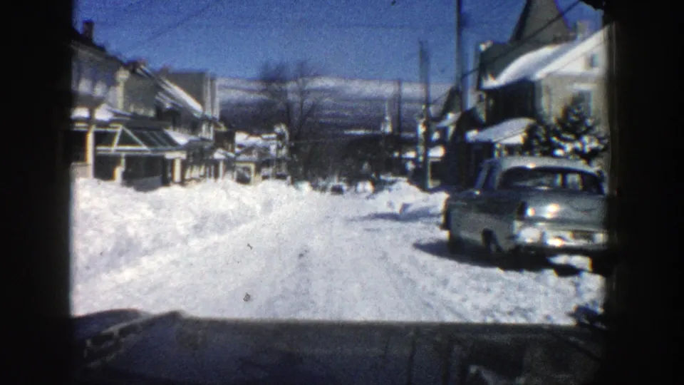 1961 Aspen winter street with piled snow, a parked mid-century car, houses, bare trees, and a moving-vehicle viewpoint.