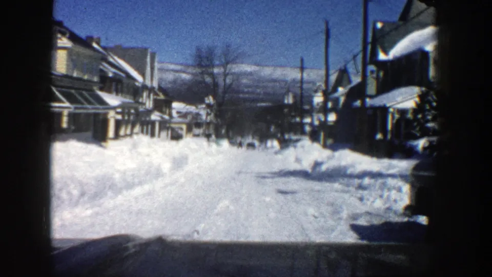 Aspen residential road in winter, filmed from inside a vehicle with snowbanks, houses, utility poles, and distant hills visible.