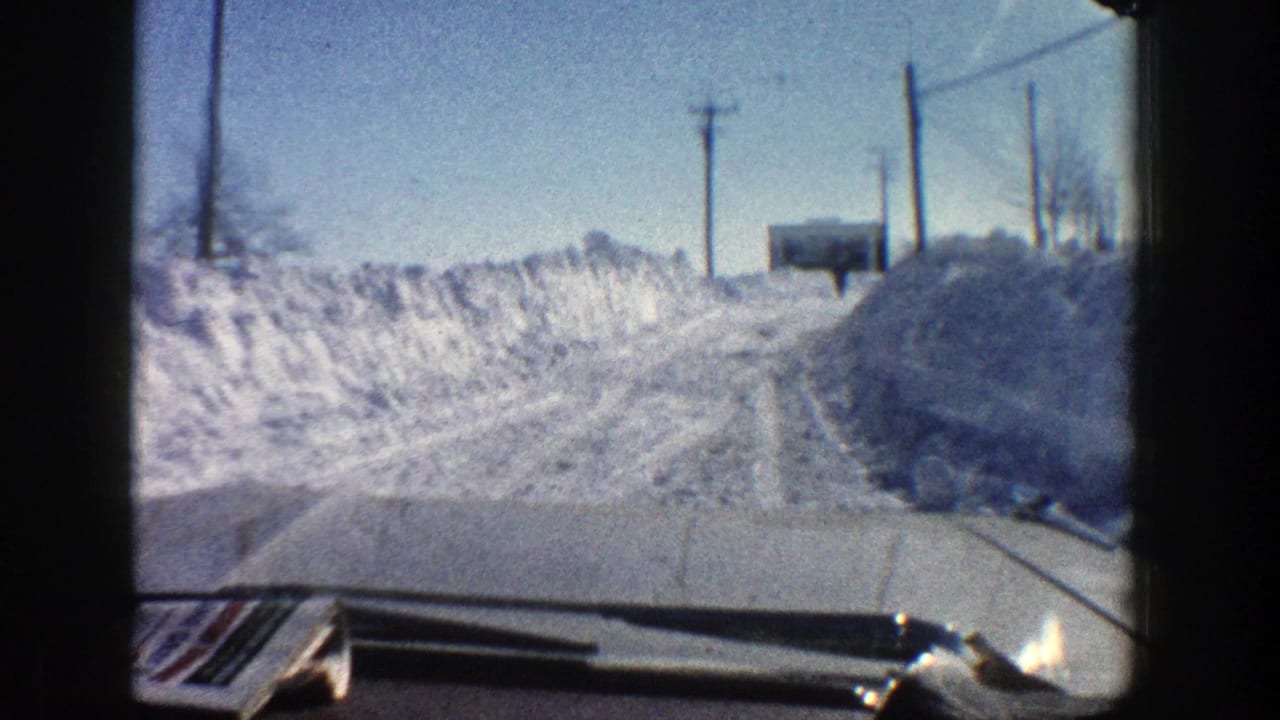 1961 Aspen, Colorado winter road view from inside a moving car, with deep snowbanks, utility poles, and the car hood visible.