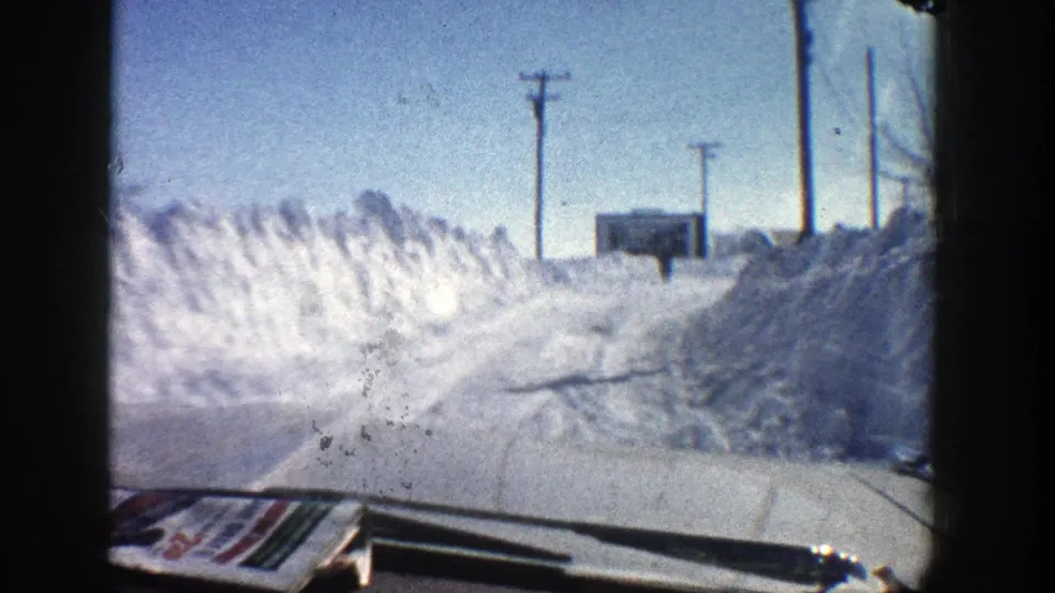 1961 Aspen, Colorado home-movie frame from inside a car approaching a snowbank-lined road with utility poles and a small structure ahead.