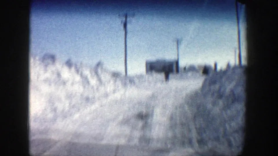 Motion-blurred 1961 Aspen, Colorado winter driving view with snowbanks, utility poles, a roadside structure, and a distant possible pedestrian.