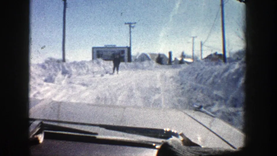 1961 Aspen, Colorado winter road scene from inside a car, showing deep snowbanks, utility poles, a small structure, and a distant pedestrian.