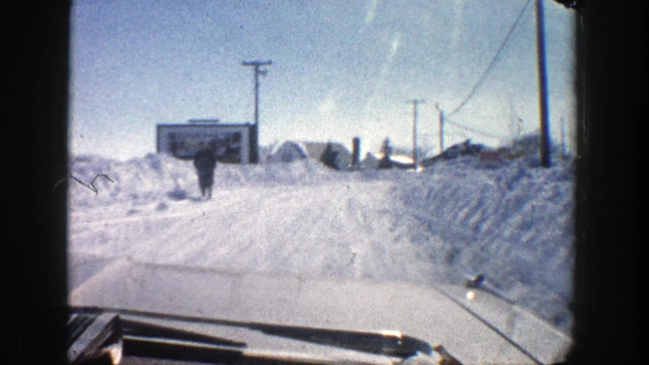 Snowy road seen through a car windshield with a distant pedestrian and roadside snowbanks.