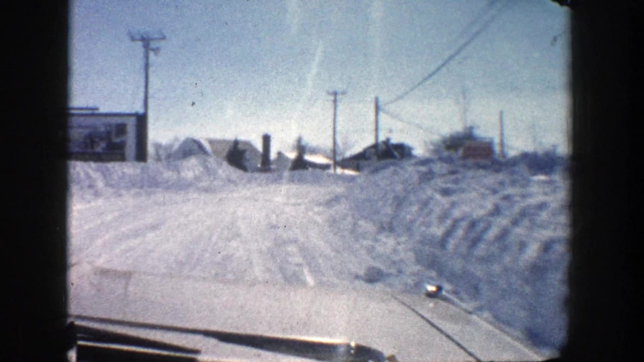 Forward car view of a snowy road with high snowbanks, poles, wires, and distant buildings.