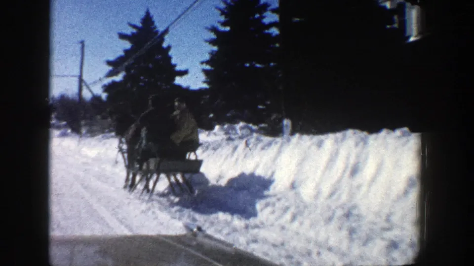 1961 Aspen car POV captures a horse-drawn sleigh with riders passing close by amid snowbanks, trees, and winter road shadows.