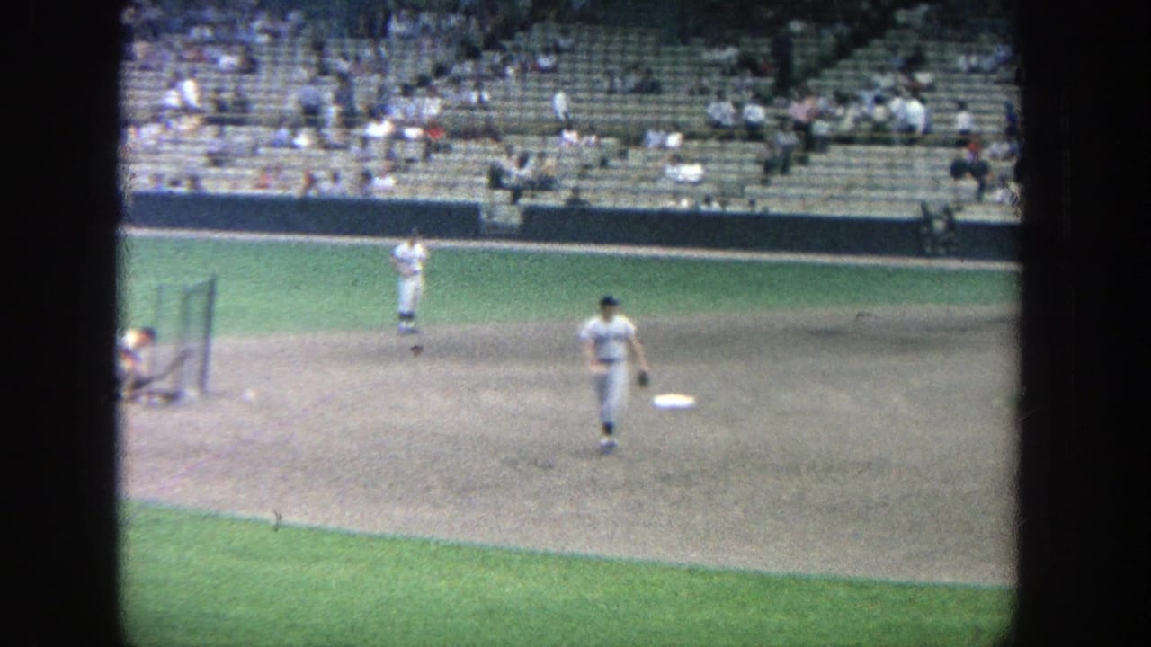 Distant baseball players warming up on the infield with spectators in stadium seats.