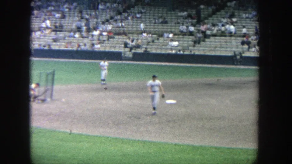1964 Yankee Stadium home-movie frame showing players in white uniforms warming up near the infield as spectators sit in the stands.