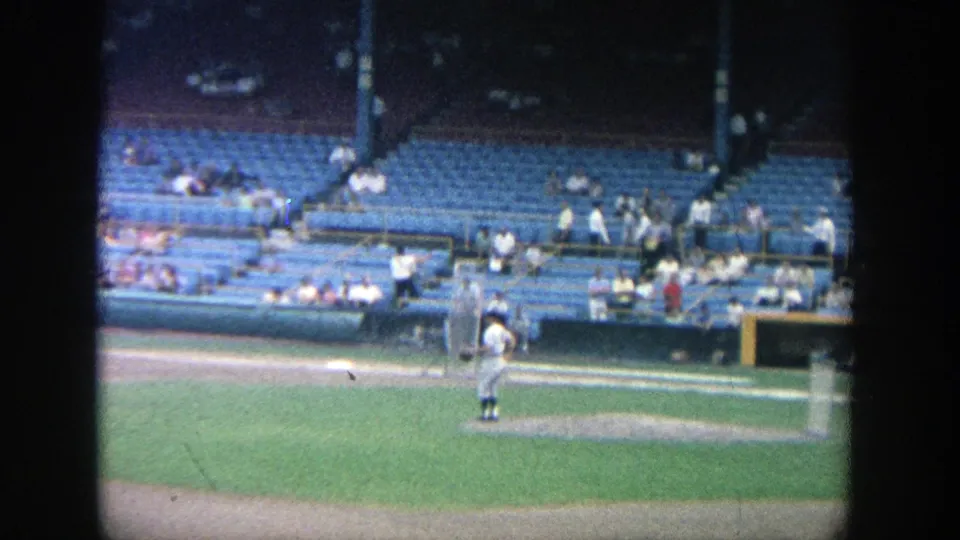 1964 Yankee Stadium home-movie frame showing a player near the mound or practice area, with blue seating and scattered spectators in the background.