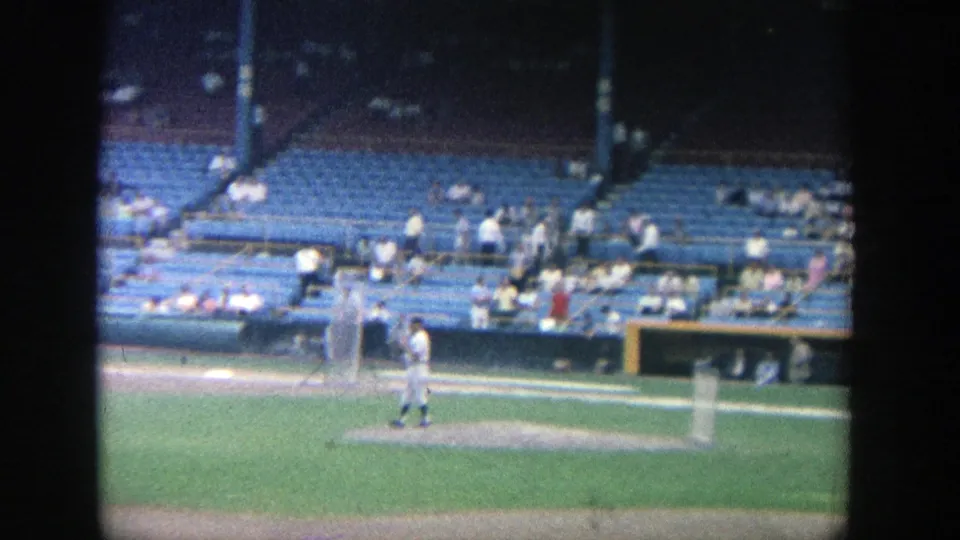 Distant 1964 Yankee Stadium warmup view with a uniformed player, practice screen, green field, and spectators spread across blue seats.