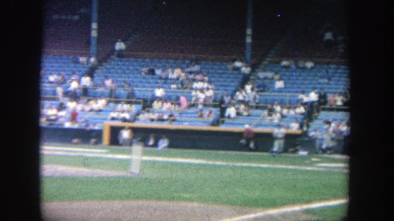 Baseball field viewed from the stands with blue seats and scattered spectators behind the play area.
