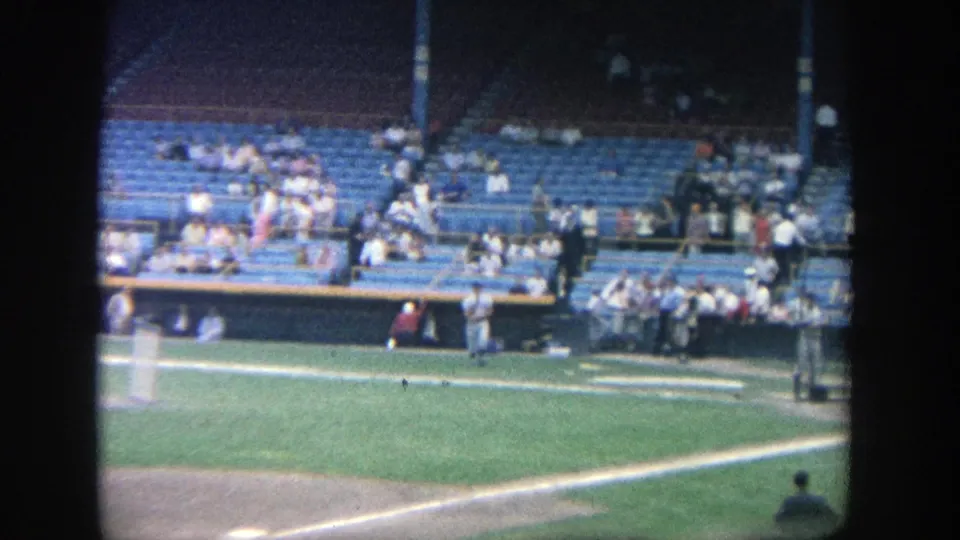 1964 Yankee Stadium home-movie frame showing the field edge and grandstand, with spectators spread across blue seats.