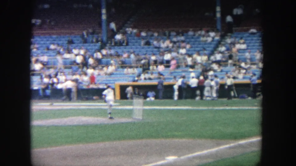 1964 Yankee Stadium 8mm frame with a pitcher on the mound, field grass, dugout area, and spectators in blue seats.