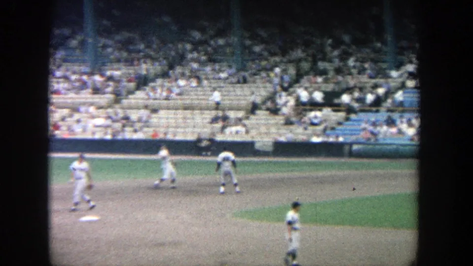 1964 Yankee Stadium frame showing infielders in white uniforms and a sparse crowd in the stands beyond the field.