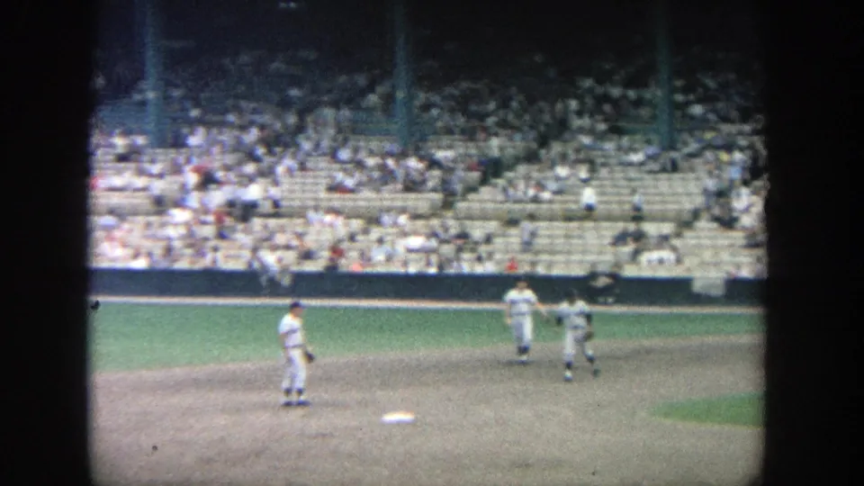 Home-movie frame from Yankee Stadium in 1964 showing players near the infield and scattered spectators behind them.