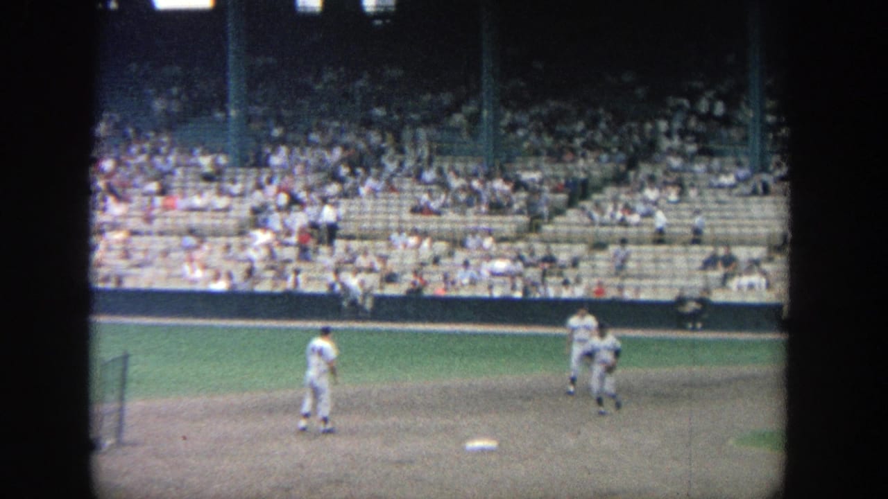 1964 Yankee Stadium home-movie frame showing baseball players near the infield with spectators seated in the stands.