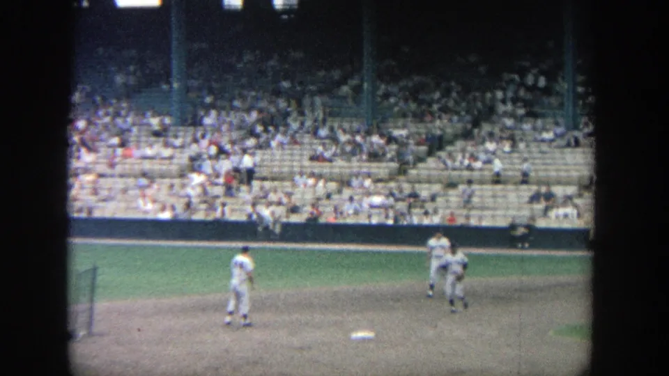 1964 Yankee Stadium home-movie frame showing baseball players near the infield with spectators seated in the stands.