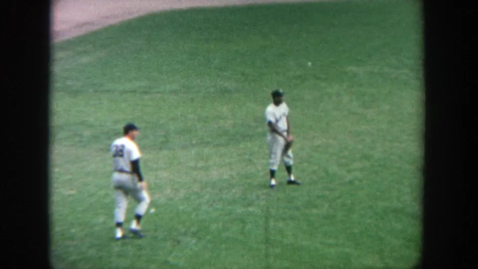 1964 Yankee Stadium home-movie frame with two white-uniformed baseball players spaced apart on the green outfield.
