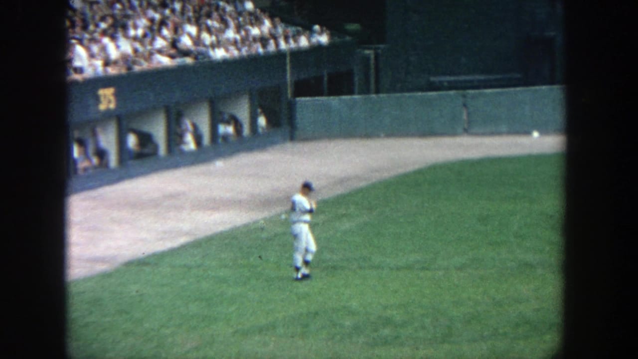 Home-movie view of a uniformed baseball player from behind on the Yankee Stadium outfield, with stands and wall visible.
