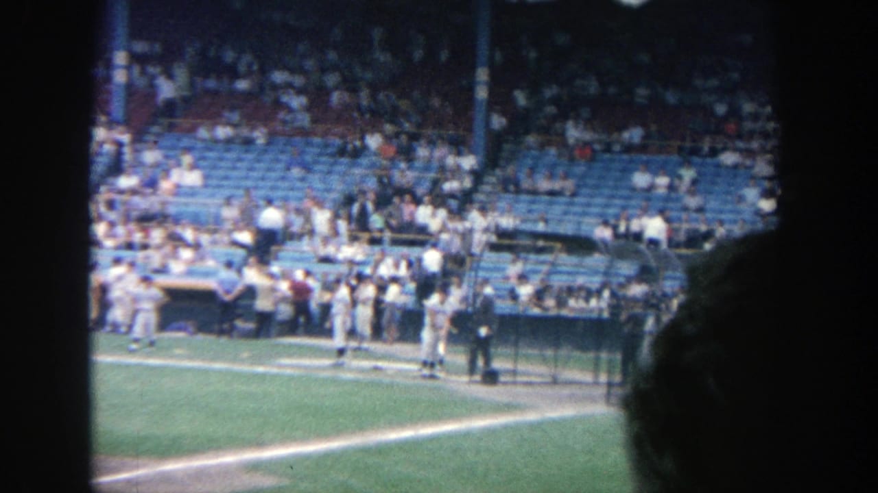 Baseball players practice near the dugout with blue stadium seats and spectators behind them.