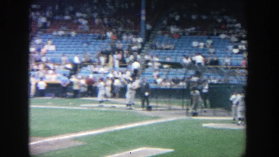 Wide 8mm frame of Yankee Stadium practice, showing a player near home plate, teammates by the dugout, and fans behind them.