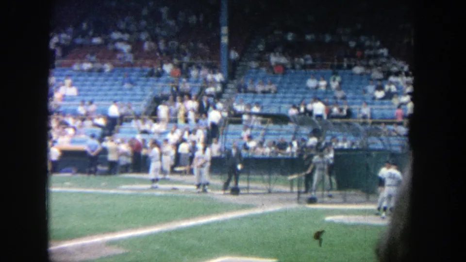 1964 Yankee Stadium practice scene with several uniformed players, dugout railings, spectators, and a dark edge from the home-movie framing.