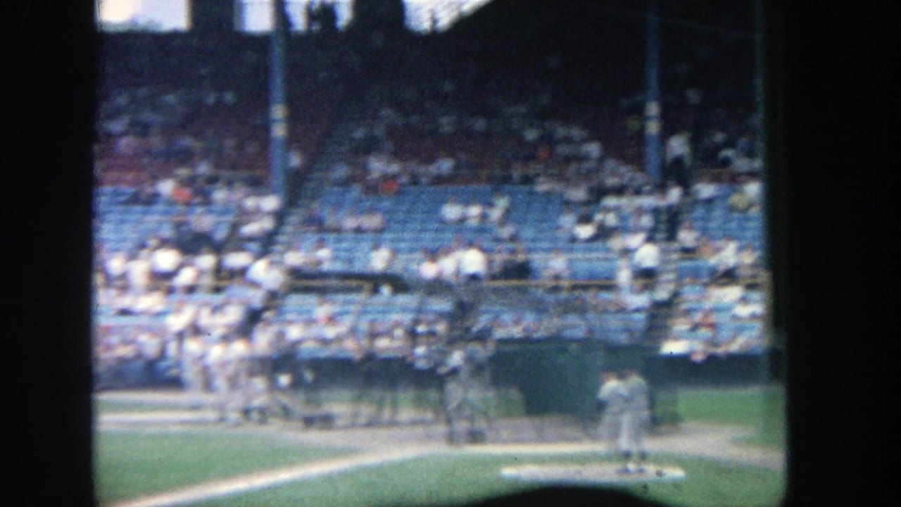 Batter stands near the plate while players and spectators appear in the background.