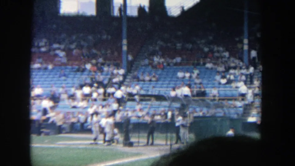 Amateur 8mm frame from Yankee Stadium showing players clustered by the batting area and fans in blue seats.