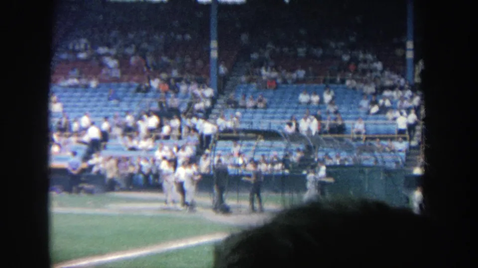 1964 Yankee Stadium home-movie still with players near the field edge, a cage structure, and fans in blue seating.