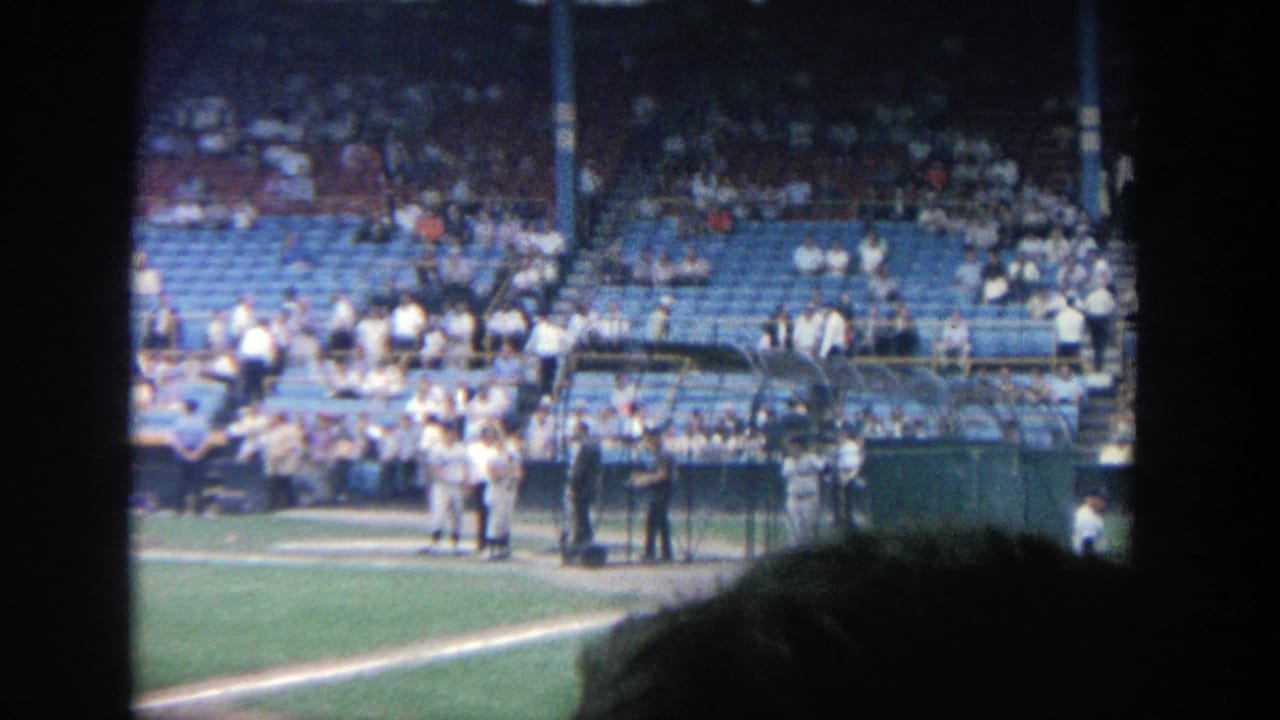 1964 Yankee Stadium home-movie view of players near the plate area with blue stands and spectators in the background.