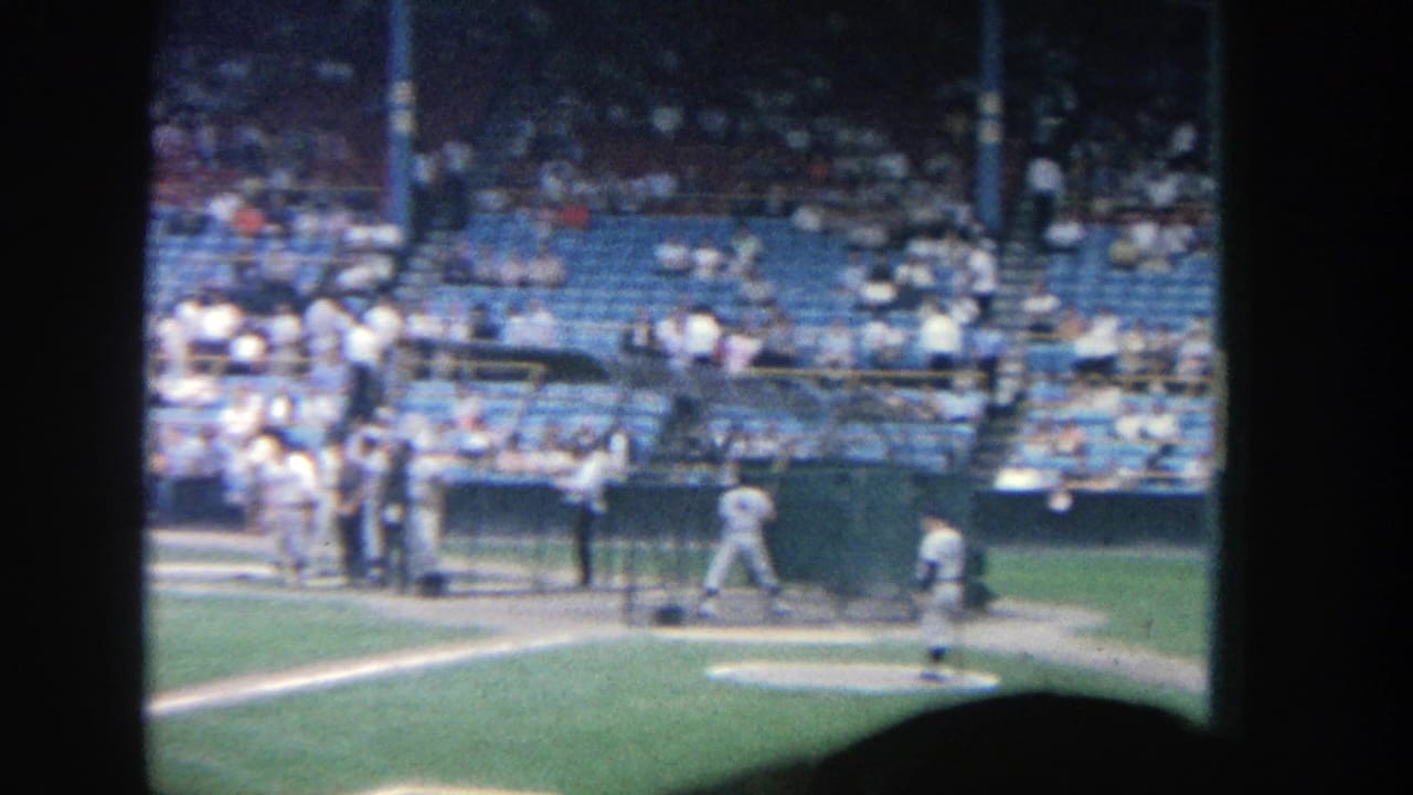 Pitcher on the mound with players near home plate and spectators in the stands.